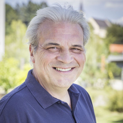 Ein lächelnder Mann in einem blauen Poloshirt steht im Freien.