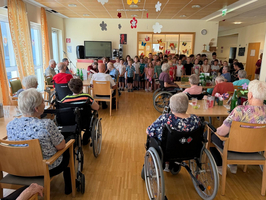 Eine Gruppe von Kindern steht zur Aufführung vor einer versammelten Menge älterer Menschen in einem Gemeinschaftsraum. Einige Senioren sitzen in Rollstühlen, während andere auf Stühlen sitzen. Die Atmosphäre scheint freundlich und fröhlich zu sein.