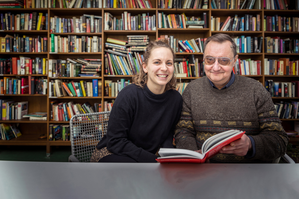 WohnenPlus Riga - zwei Bewohner:innen sitzen an Tisch vor Bücherregal und lesen in Buch