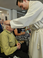 Ein junger Priester segnet einen älteren Mann in einem Stuhl.