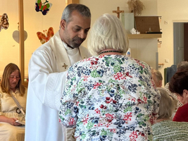 Ein Priester in weißem Gewand steht vor einer älteren Frau mit blumigem Oberteil. Er reicht ihr etwas, während eine Gruppe von Personen im Hintergrund sitzt und zuschaut. Die Szene spielt sich in einem hellen Raum ab, der gemütlich wirkt.