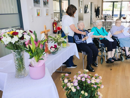 Eine Pflegekraft steht neben drei Seniorinnen, die in Rollstühlen sitzen und Liederblätter halten. Auf einem Tisch sind Blumenarrangements und ein Kreuz platziert. Der Raum ist hell und freundlich gestaltet.