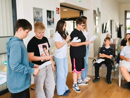 Eine Gruppe von vier Jugendlichen steht in einem Raum und hält Blätter in den Händen. Sie sprechen miteinander, während andere Schüler im Hintergrund aufmerksam zuhören. Die Atmosphäre wirkt freundlich und informell.