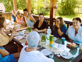 Eine Gruppe von Frauen sitzt an einem Tisch im Freien, umgeben von Bäumen. Sie lachen und unterhalten sich, während sie Getränke und Snacks genießen. Der Tisch ist gedeckt mit Gläsern, Flaschen und Tellern. Sonnenschein schafft eine entspannte Atmosphäre.