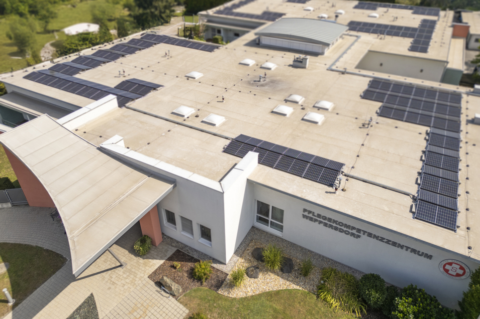 Dachansicht eines Pflegekompetenzzentrums mit mehreren Solarpanelen. Die Umgebung ist mit Grünanlagen gestaltet.