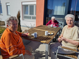 Drei ältere Frauen sitzen bei sonnigem Wetter an einem Tisch im Freien.
