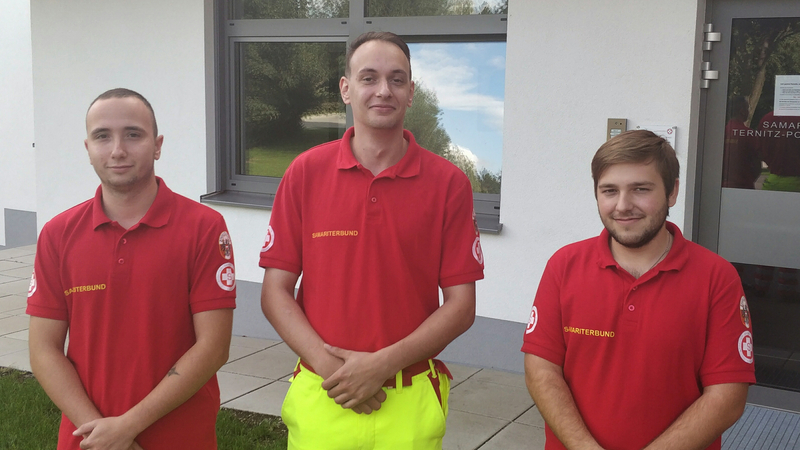 Drei Rettungssanitäter in roten Uniformen stehen lächelnd vor einem Gebäude.