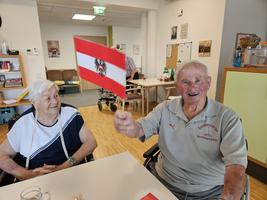 Ein älterer Mann sitzt an einem Tisch und hält eine kleine Österreich-Flagge hoch. Neben ihm sitzt eine ältere Frau, die ihn anlächelt. Im Hintergrund sind weitere Personen und eine helle, freundliche Umgebung zu sehen.