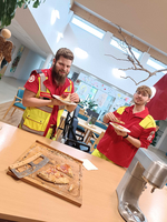 Zwei Mitarbeiter in roten und gelben Uniformen stehen in einem hellen Raum mit Möbeln und Pflanzen. Sie halten Teller mit Essen in den Händen. Auf einem Tisch liegt ein angeschnittenes Stück Pizza. Im Hintergrund sind einige Kunstwerke zu sehen.