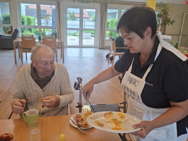 Eine Pflegerin serviert einem älteren Mann Essen am Tisch.