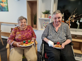 Zwei ältere Frauen sitzen in einem heller Raum und halten Teller mit Essen. Die Frau links trägt ein buntes Oberteil und lächelt, während die Frau rechts, die im Rollstuhl sitzt, ebenfalls lächelt und eine Bandage an ihrer Hand hat. Im Hintergrund sind dekorative Pflanzen sichtbar.
