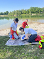 Drei Rettungskräfte am Ufer eines Gewässers kümmern sich um eine Person, die auf einer Decke liegt. Zwei Helfer beugen sich über die Person, während der dritte im Hintergrund aufrecht steht. Alle tragen Schutzkleidung und wirken konzentriert.