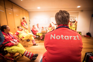 Eine Gruppe von Rettungskräften in einem Raum, die in roten und gelben Uniformen gekleidet sind. Eine Person mit dem Rücken zur Kamera trägt eine Jacke mit der Aufschrift 