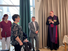 Eine Gruppe von Personen steht in einem Raum mit einem Tisch, der religiöse Gegenstände zeigt. Eine Frau spricht lächelnd zu einem Mann in einem Anzug, während ein Bischof in liturgischer Kleidung aufmerksam zuhört. Im Hintergrund sind Vorhänge und Fenster sichtbar.