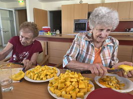 Zwei ältere Frauen schneiden und schälen Obst in einer Küchenumgebung.