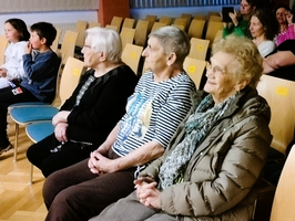 Drei ältere Frauen sitzen in einem Auditorium auf Stühlen und schauen aufmerksam. Im Hintergrund sind einige jüngere Zuschauer sichtbar. Die Frauen tragen unterschiedliche Kleidung und haben verschiedene Frisuren. Die Umgebung ist hell erleuchtet.