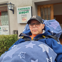 Ein Mann sitzt im Rollstuhl vor einem Gasthaus mit einem blau gepunkteten Decke.
