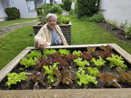 Eine ältere Frau sitzt an einem Hochbeet mit verschiedenen Salatsorten. Sie trägt einen cardigan und lächelt, während sie mit einer Hand auf die Pflanzen zeigt. Im Hintergrund sieht man eine grüne Wiese und einige Gartenmöbel.