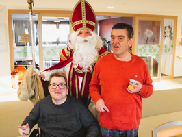Der Nikolaus steht zwischen zwei lächelnden Personen in einem hellen Raum.