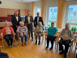 Eine Gruppe von acht Personen sitzt und steht in einem hellen Raum. Im Vordergrund sind sechs ältere Frauen auf Stühlen zu sehen, die freundlich lächeln. Im Hintergrund stehen zwei Männer in Anzügen. Der Raum ist freundlich eingerichtet, mit großen Fenstern und einer modernen Küchenecke.