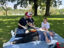 Drei Personen auf einer Wiese geben Erste Hilfe. Zwei Jugendliche im Vordergrund kümmern sich um eine Person, die auf einem blauen Tuch liegt, während eine dritte Person daneben sitzt und beobachtet. Im Hintergrund ist ein Gewässer und Bäume sichtbar.