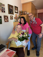 Eine ältere Frau sitzt auf einem Stuhl und hält einen Blumenstrauß in einer Vase. Neben ihr stehen zwei Personen, die sie umarmen. Im Hintergrund sind Familienfotos an der Wand sichtbar. Die Szene strahlt Freude und Zuneigung aus.
