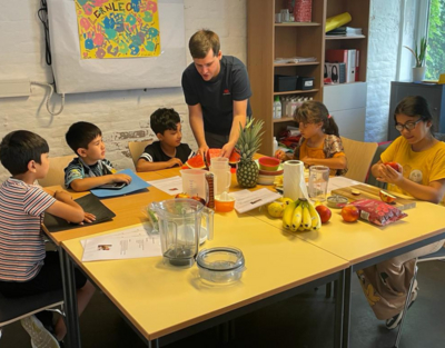 Gruppe von Kindern sitzt rund um Tisch, Mann zeigt Obst