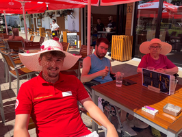 Drei Personen sitzen an einem Tisch im Freien unter Sonnenschirmen. Eine Person trägt einen großen, bunten Sonnenhut und ein rodes T-Shirt. Eine andere Person in einem blauen Tanktop hat ein Getränk in der Hand. Die dritte Person trägt einen ebenfalls großen Hut und schaut in die Kamera.