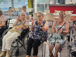 Drei ältere Frauen sitzen in Rollstühlen und applaudieren. Sie tragen bunte Kleidung und haben freundliche Ausdrücke. Im Hintergrund sind weitere Personen und ein heller, einladender Raum zu erkennen.