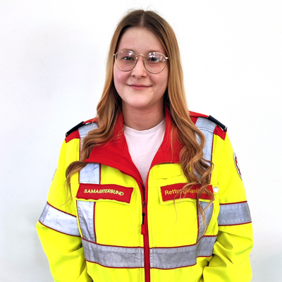 Junge Frau in gelb-roter Rettungsdienstuniform steht vor weißem Hintergrund.