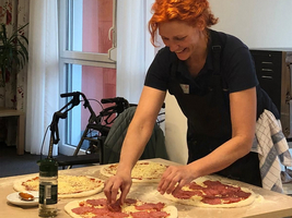 Eine lächelnde Frau mit roten Haaren bereitet Pizzaböden vor, indem sie Belag auf diese legt. Sie trägt eine Schürze und steht an einem Tisch in einem hellen Raum. Im Hintergrund ist ein Rollstuhl sichtbar und auf dem Tisch steht eine Flasche.