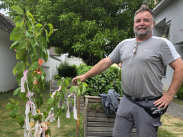 Ein Mann steht neben einem kleinen Baum, der mit bunten Bändern geschmückt ist. Er trägt ein graues T-Shirt und eine Hose, die zum Arbeiten geeignet ist. Im Hintergrund sind Bäume und ein einfacher Gartenbereich sichtbar.