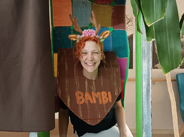 Eine fröhliche Person mit rotem, lockigem Haar trägt ein Kostüm mit einem Schild, auf dem „BAMBI“ steht. Sie hat Geweih-Nachbildungen auf dem Kopf und steht in einem bunten, handgemachten Rahmen mit verschiedenen dekorativen Elementen.