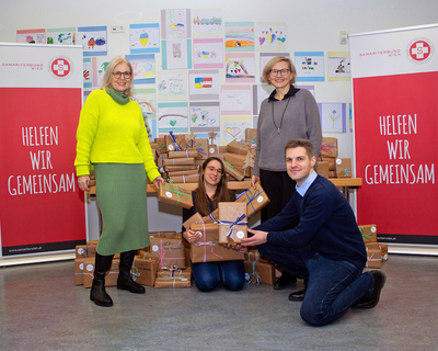 Gruppenfoto Geschenkübergabe, Susanne Drapalik und Mitarbeiter:innen von Expleo in Ukraine Sozialberatung, viele Geschenke im Hintergrund, rechts und links Rollups Samariterbund Wien Helfen wir gemeinsam, im Hintergrund Kinderzeichnungen