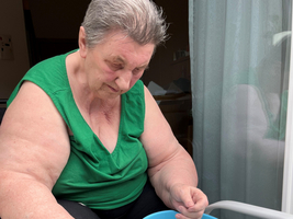 Eine ältere Frau mit kurzen grauen Haaren sitzt in einem Stuhl und sortiert herausgerissene Strohhalme auf dem Boden. Sie trägt ein grünes Oberteil und hat eine blaue Schüssel vor sich, in die sie die Strohhalme legt. Der Hintergrund zeigt einen Raum mit Fenster und Vorhängen.