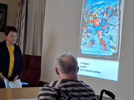 Eine Präsentation in einem Raum, wo eine Frau steht und mit einer Gruppe von Menschen spricht. Auf einer Leinwand ist ein farbiges Bild zu sehen, das eine Szenenbeschreibung enthält. Eine Person im Rollstuhl sitzt in der Frontansicht.