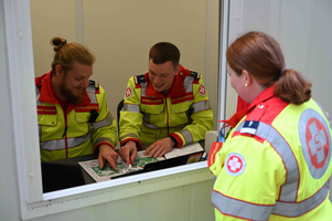 Drei Rettungsdienstmitarbeiter in leuchtend gelben Uniformen sitzen an einem Tisch und schauen auf einen Plan. Zwei von ihnen zeigen auf eine Stelle auf dem Plan, während eine dritte Person im Hintergrund steht und zuschaut.