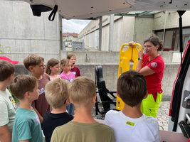 Eine Ausbilderin zeigt Kindern eine gelbe Trage in einem medizinischen Umfeld.