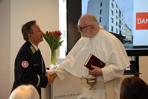 Zwei Männer schütteln sich die Hände. Der Mann links trägt eine Uniform mit Abzeichen, während der Mann rechts einen weißen Umhang und eine Brille trägt. Im Hintergrund sind ein Bildschirm mit einem Gebäude und eine Vase mit roten Tulpen sichtbar.