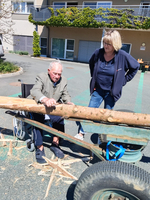 Ein älterer Mann sitzt im Rollstuhl und bearbeitet einen Holzstamm auf einem Wagen. Eine Frau steht neben ihm und beobachtet ihn. Im Hintergrund sind Gebäude und Bäume zu sehen. Der Ort wirkt freundlich und gut beleuchtet.