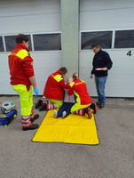 Drei Sanitäter führen eine Übung durch. Die Übungspuppe lehnt an einer Garagenwand.