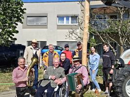 Eine fröhliche Gruppe von Menschen, darunter ein älterer Herr im Rollstuhl, posiert vor einem Gebäude. Einige Personen halten Musikinstrumente wie ein Tuba und ein Akkordeon. Sie stehen in einem Gartenbereich mit ein paar Bäumen und einem Traktor.