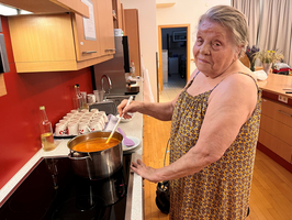 Eine ältere Frau in einem bunten Kleid steht in einer modernen Küche und rührt in einem großen Topf mit Suppe. Im Hintergrund sind Küchenschränke und eine Reihe von Tassen zu sehen. Sie schaut freundlich zur Kamera.