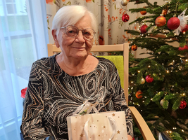 Ältere Frau sitzt lächelnd mit Geschenk vor einem Weihnachtsbaum.