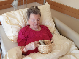 Eine ältere Person sitzt im Bett und hält einen kleinen Korb mit Gegenständen in den Händen. Sie trägt ein rotes Oberteil und schaut nachdenklich. Der Hintergrund zeigt eine Krankenhausumgebung mit Kissen und Bettwäsche.