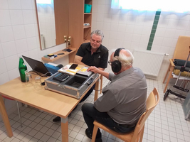 Zwei Männer sitzen an einem Tisch mit elektronischen Geräten und einem Koffer.