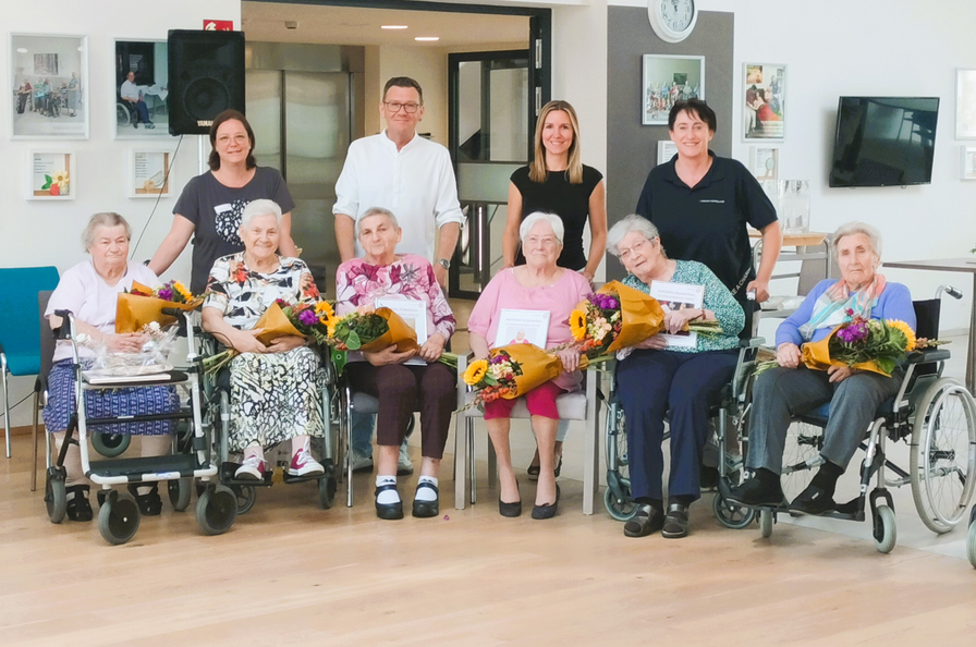 Feierliche Gruppe von Senioren mit Blumenstraußen und Urkunden in einem Wohnheim.
