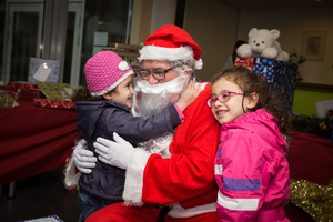 Ein fröhlicher Weihnachtsmann umarmt zwei lächelnde Kinder in festlicher Umgebung.