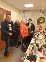 Eine Gruppe von fünf Personen steht in einem Raum und betrachtet weihnachtliche Dekorationen auf einem Tisch. Sie lächeln und sind in festlicher Kleidung, während sie sich um einen Tisch mit verschiedenen dekorativen Gegenständen versammeln, darunter Kerzen und kleine Figuren.