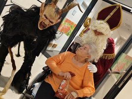 Eine ältere Frau im Rollstuhl wird von einem Krampus und Nikolaus besucht.